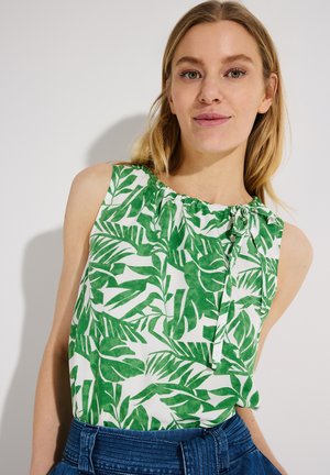 Frau trägt ärmelloses weißes Oberteil mit grünem tropischem Blattdruck und blaue hoch taillierte Jeans, lächelt vor einfachem Hintergrund.