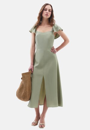 Abito midi verde salvia con maniche a volant e spacco frontale, abbinato a una borsa intrecciata e sandali dorati con cinturini. Tessuto dalla texture liscia.