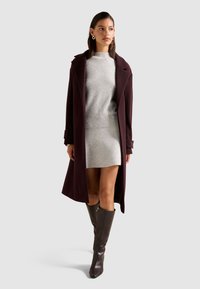 Abrigo largo burdeos con solapas amplias, combinado con un suéter gris claro y una falda a juego. Botas altas de cuero marrón completan el look.