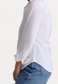 Blusa blanca de manga larga con un cuello alto, puños abotonados y un dobladillo ligeramente curvado, combinada con jeans azules. Tejido suave y ligero.