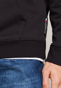 Sudadera negra con acabado texturizado, que presenta una pequeña etiqueta con el logo de una bandera y puños acanalados. Combinada con jeans de mezclilla azules.