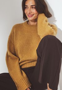 Mosterdgele gebreide trui met een geribbelde ronde hals en brede mouwen, gecombineerd met donkere broek, tegen een lichte achtergrond.