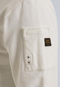 Biely bavlnený sveter s rukávmi, ktorý má náprsné vrecko so zdvojenými kovovými očkami a značkovou etiketou. Hladká textúra a ležérny dizajn.