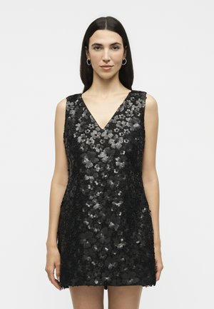 Γυναίκα με ίσια σκούρα μαλλιά φοράει αμάνικο μαύρο φόρεμα διακοσμημένο με παγιέτες σε λουλουδένια σχέδια, στέκεται σε λευκό φόντο.