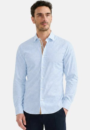 Uomo con una camicia azzurro chiaro a maniche lunghe con colletto e pantaloni scuri, in piedi con una mano in tasca.