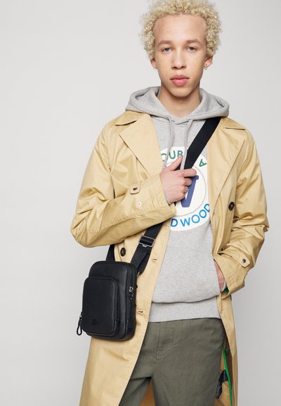 Sort læder crossbody taske med en tekstureret overflade, lynlåslomme og justerbar rem, båret over en grå hættetrøje og en beige trench coat.
