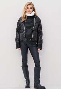 Fekete steppelt puffer dzseki fehér díszítéssel, dupla gombos záródással és magas gallérral, fekete fényes leggings-szel és térdig érő csizmával párosítva.