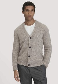 Lichtbruine cardigan van textuur gebreid, met een v-hals, vier knopen aan de voorkant en geribde boorden en onderkant. Draagt over een wit T-shirt.