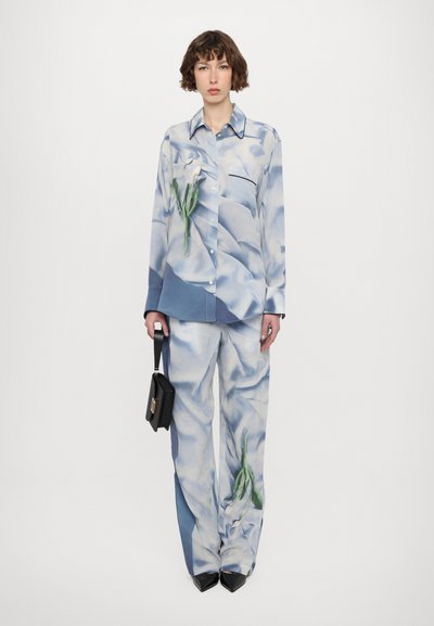 Floral-patterned silk shirt and matching trousers in light blue hues, featuring a green plant design. Black handbag and pointed black shoes.