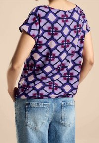 Top a maniche corte viola con un motivo a diamante in tonalità di rosa e rosso scuro, abbinato a jeans azzurri chiari. Tessuto morbido con una vestibilità rilassata.