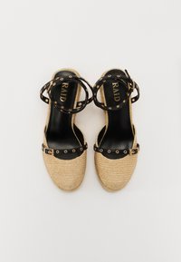 Wedges de esparto tejidos en marrón con correas negras en los tobillos que cuentan con tachuelas circulares y un detalle de hebilla. Forro interior negro y etiqueta de marca visible.