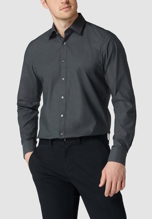 Homme portant une chemise cintrée gris foncé à boutons avec manches longues et un pantalon noir sur un fond uni.