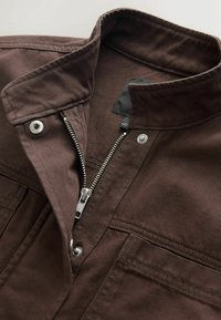 Veste marron avec fermeture éclair métallique partiellement ouverte, boutons-pression au col et à la poche poitrine, détails de couture visibles.