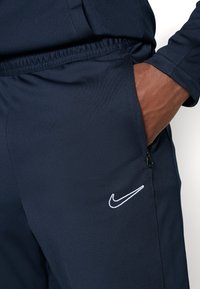 Marineblauwe sportieve joggers gemaakt van gestructureerde stof, met een wit Nike-logo aan de linkerkant en een ritszak aan de rechterkant.