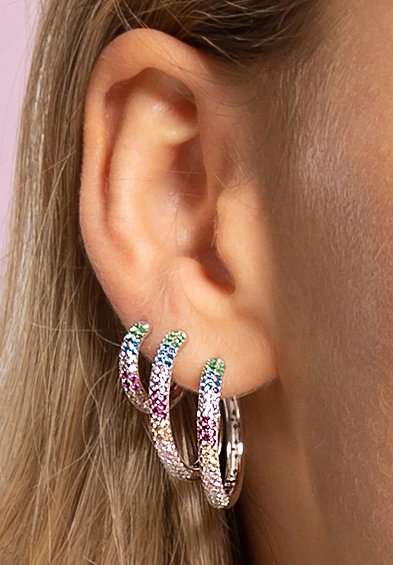 Trois boucles d'oreilles en anneau entrelacées en ton argenté, ornées d'un dégradé de strass pastel en vert, rose et bleu, posées contre une oreille.