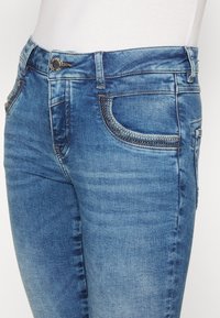 Närbild på blå denimjeans med silverdetaljer på framfickorna och en metallknäppning, som bärs med en vit topp.