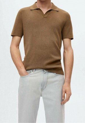 Mann trägt braunes Poloshirt mit kurzen Ärmeln und hellgraue Jeans, eine Hand in der Tasche, neutraler Hintergrund.