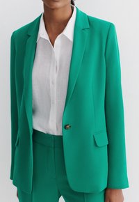Blazer verde su misura con una texture liscia, singolo bottone dorato, rever a punta e tasche frontali a patta, indossato sopra una camicia bianca con bottoni.