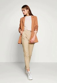 Γυναίκα που φοράει ταμπά σακάκι πάνω από λευκό μπλούζα και μπεζ παντελόνι, στέκεται με το ένα χέρι στην τσέπη και κοιτάζει προς τα κάτω, οι λευκές αθλητικές της παπούτσια είναι ορατές.