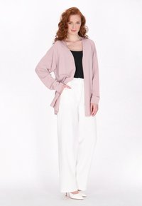 Cardigan rosa chiaro con maniche lunghe, apertura frontale e fessure laterali, abbinato a una maglietta nera e pantaloni bianchi a gamba larga. Scarpe bianche a punta visibili.