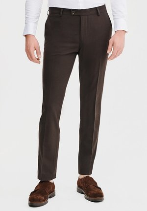 Brązowe eleganckie spodnie o prostym kroju, z płaskim przodem i szlufkami na pasek. Noszone z brązowymi zamszowymi butami i białą koszulą. Gładka tekstura.