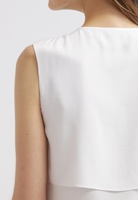 Gros plan sur l'épaule et le haut du dos d'une femme portant un haut blanc sans manches avec un détail superposé dans le dos, sur un fond uni.