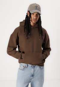 Sweatshirt castanho com bolso canguru, corte solto e punhos canelados, combinado com jeans de denim azul claro e um boné cinza.