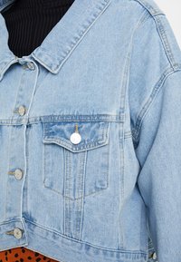 Ljusblå denimjacka med en kortad design, silverfärgad knapputrustning, två framfickor och en texturerad yta.