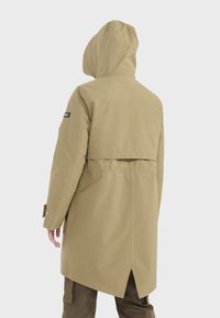 Veste imperméable beige avec capuche, dos allongé, taille élastique et poignets à bouton-pression. Ventilation arrière conçue pour la circulation de l'air.