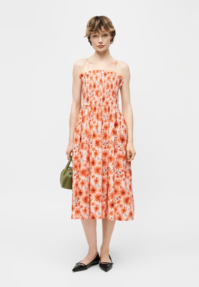 Νεαρή γυναίκα που φοράει αμάνικο, πορτοκαλί φόρεμα με λουλούδια μήκους μέχρι τη μέση της γάμπας, κρατώντας μια μικρή τσάντα σε ελιά πράσινο χρώμα και φορώντας μαύρα μυτερά φλατ παπούτσια.