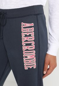 Navyblauwe trainingsbroek met verticale roze en witte "Abercrombie" logo op de dij, gedragen met een witte lange mouw shirt, hand op de heup.