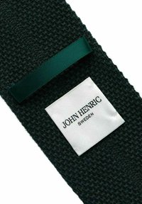 Tummanvihreä neulottu tekstiili, jossa on sileä satiinirusetti ja valkoinen etiketti, jossa lukee "JOHN HENRIC SWEDEN" mustalla tekstillä.
