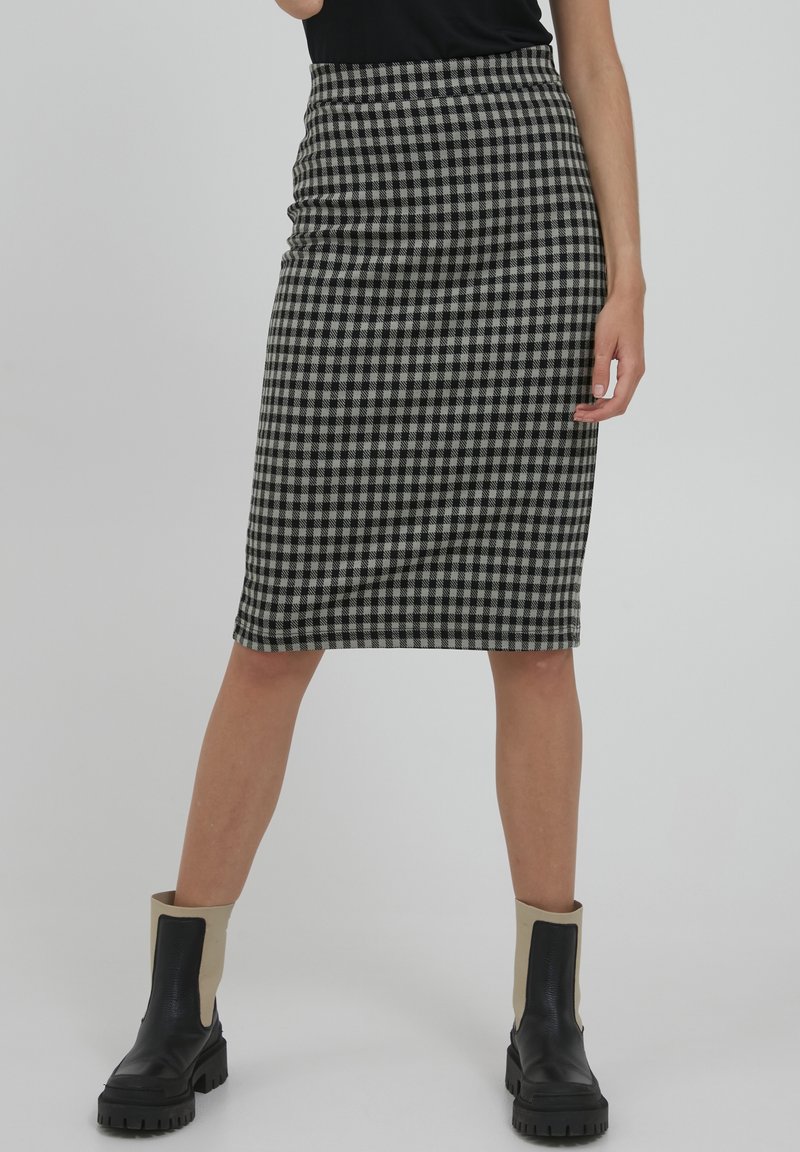 Femme portant une jupe crayon à carreaux noirs et blancs longueur genou, un haut noir et des bottes noires avec des manchettes beiges au niveau de la tige.