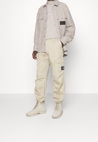 Pantaloni cargo beige con polsini elastici, abbinati a una maglietta a maniche lunghe grigio chiaro e una camicia abbottonata, tutti in tonalità neutre.