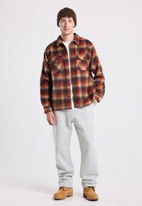 Camicia di flanella a quadri rossi, arancioni e neri, abbinata a pantaloni sportivi grigi e stivali marroni con lacci. Presenta due tasche sul petto.