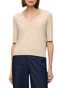 Maglietta a maniche corte in maglia beige con scollatura a V, caratterizzata da una linea verticale di piccole perforazioni al centro. Texture liscia, vestibilità casual.