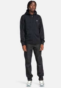 Schwarze Kapuzenjacke mit Reißverschluss aus weichem Stoff, ausgestattet mit einer Vordertasche und verstellbarem Kordelzug an der Kapuze, kombiniert mit dunklen Jeans und grauen Sneakers.