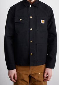 Giacca nera in tela con bottoni a scatto bronzei, due tasche sul petto e una piccola toppa con logo. Presenta un colletto in velluto a coste e un design moderno.