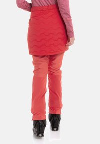 Punainen eristetty hame, jossa aaltoileva tikattu kuvio, puettuna oranssien hiihtohousujen päälle. Mustat hiihtomonot metallisilla yksityiskohdilla täydentävät asun.