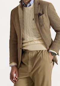 Brązowa marynarka w jodełkę, kremowy sweter w warkocze i beżowe spodnie. Widoczny niebieski kołnierzyk koszuli. Zawiera wzorzysty poszetkę i zegarek.