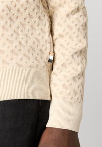 Gros plan sur la manche d'un pull beige texturé avec un poignet côtelé et une petite étiquette en tissu tricolore sur un pantalon noir.