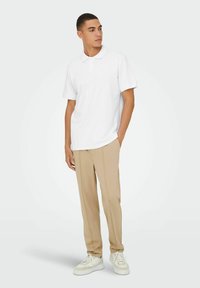 Hvid polo skjorte med klassisk krave, korte ærmer, parret med beige bukser og hvide sneakers, stående foran en neutral baggrund.