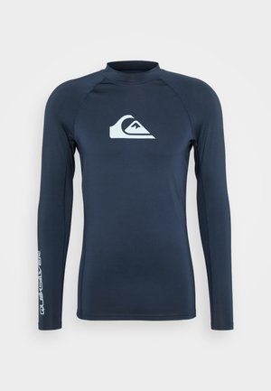 Marineblaue Langarm-Rashguard aus dehnbarem Material, mit einem weißen Logo auf der Brust und Text entlang des linken Ärmels. Glatte Textur.