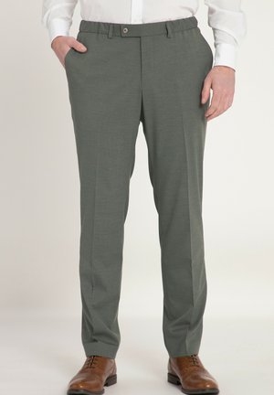 Uomo con pantaloni da abito grigi, camicia bianca e scarpe eleganti in pelle marrone, con le mani parzialmente nelle tasche, in piedi su sfondo bianco.