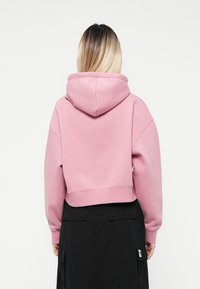 Hoodie rosa tagliato con cappuccio regolabile, maniche oversize e una texture morbida. Indossato sopra una gonna cargo nera.