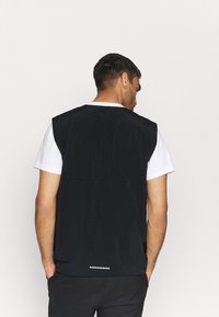 Homem de costas, vestido com um colete preto sem mangas sobre uma camisola branca e calças pretas, em pé contra um fundo branco simples.