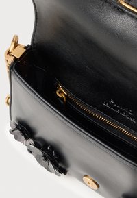 Bolsa de mão em couro preto com acabamentos em dourado, aba aberta revelando um bolso interior com fecho, e embellishments florais pretos decorativos na parte da frente.