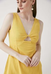 Gelbes Kleid mit tiefem V-Ausschnitt und verdrehtem Detail an der Brust. Hergestellt aus strukturiertem Stoff, mit klaren Linien und einem fließenden Schnitt.