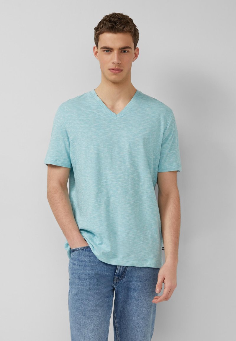 Młody mężczyzna w jasnoniebieskim T-shircie z krótkim rękawem i dekoltem w szpic oraz niebieskich dżinsach, stojący na tle jednolitego jasnoszarego tła.