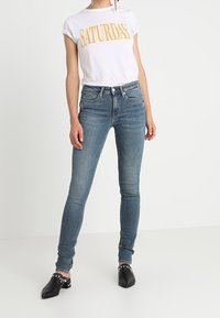 Camiseta blanca con la palabra "SÁBADO" en texto amarillo, combinada con jeans de mezclilla azul de corte ajustado. Se lleva con calzado negro con tachuelas blancas.